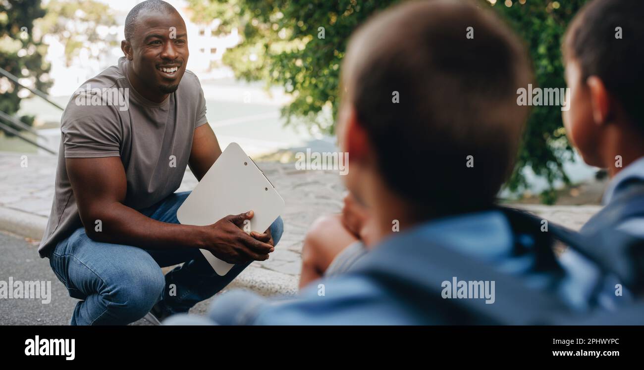 Teacher talking to his students outside class in a primary school. Elementary school educator motivating a group of children. Academic mentorship and Stock Photo