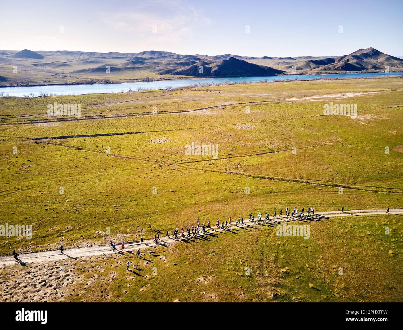 Aerial drone view of crowd people running competition down dusty country road near river and hills at sunset. Trail running at the steppe near Ili riv Stock Photo