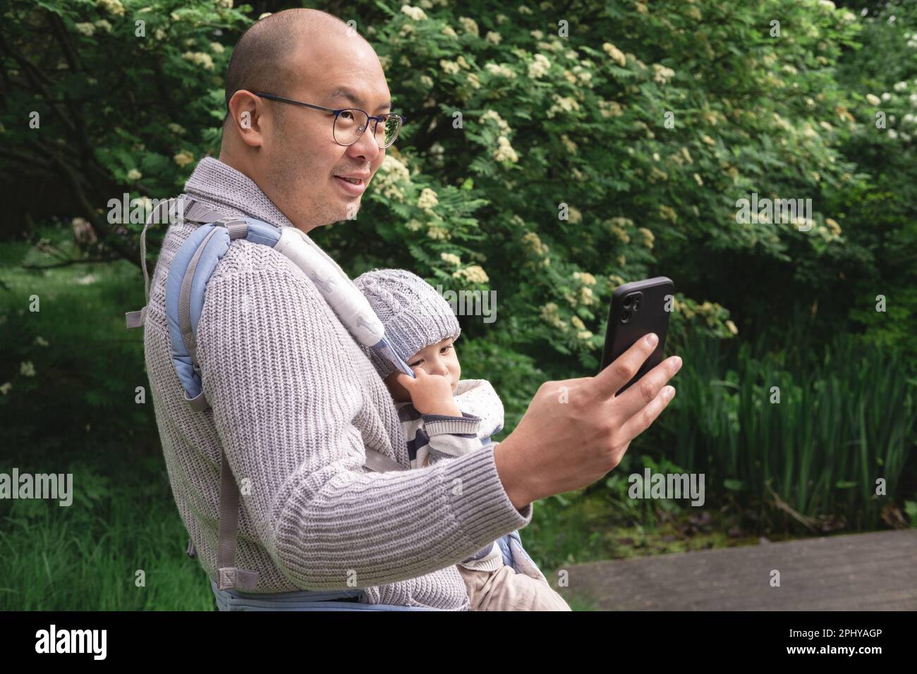 A smiley new father taking a photo with his cell phone while carrying his male infant with a baby carrier in nature at the park in spring season. Brit Stock Photo