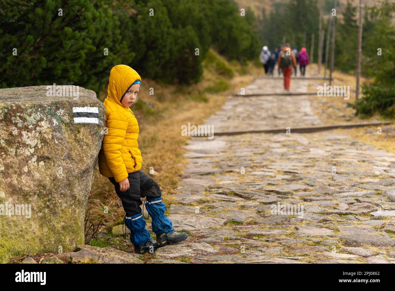 The child rests on the mountain trail, standing leaning against a large boulder. Tourists are marching in the background. Polish mountains Stock Photo