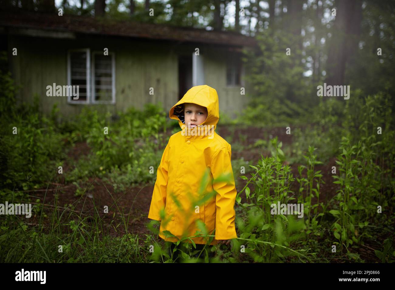 A child in a yellow coat standing in the woods in front of an old wooden house. A six-year-old boy, Poland, Europe Stock Photo