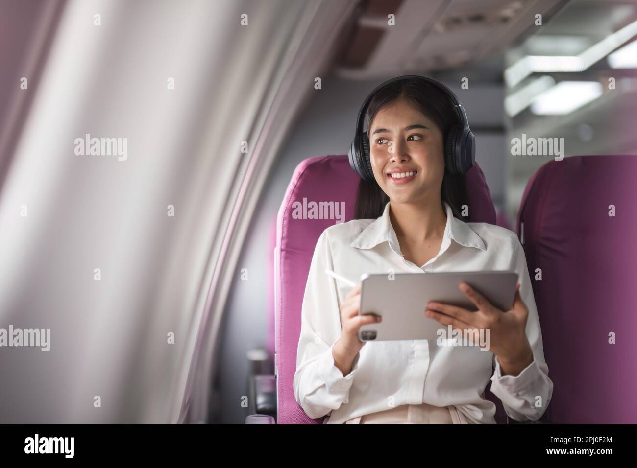 Cheerful female passenger in headphones for noise cancellation watching online movie during intercontinental flight in cabin of aircraft, happy young Stock Photo