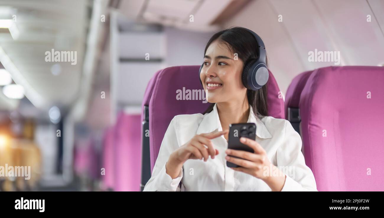 Cheerful female passenger in headphones for noise cancellation watching online movie during intercontinental flight in cabin of aircraft, happy young Stock Photo