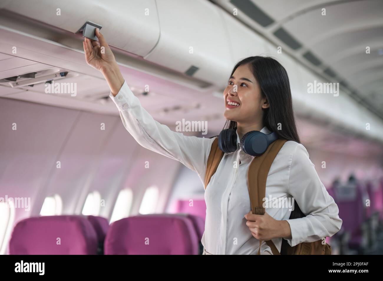 Young asian woman travel by airplane. Passenger putting hand baggage in lockers above seats of plane Stock Photo