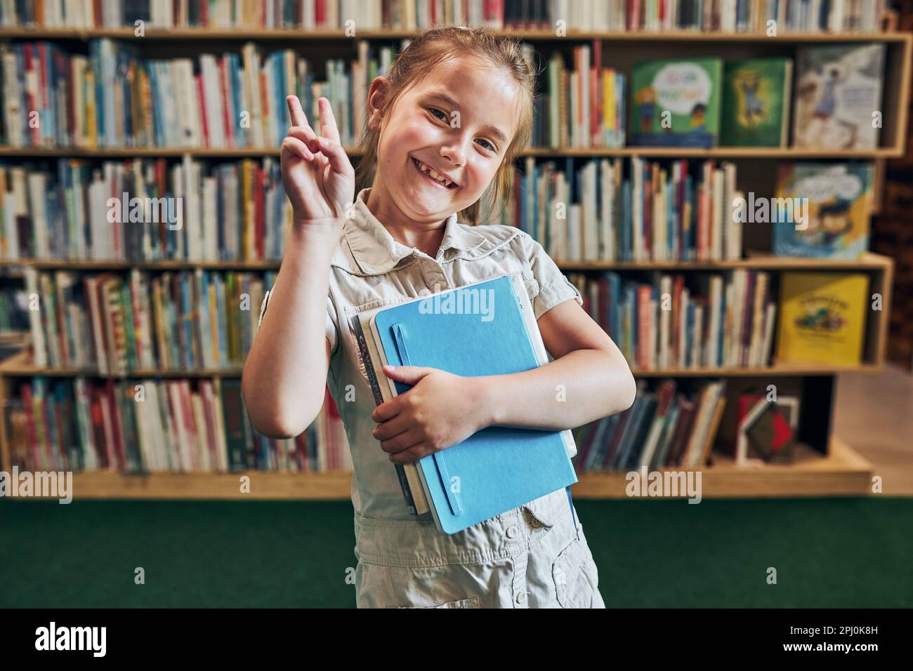 Back to school. Happy little girl at school again. Smiling student making V sign hand gesture holding books standing at front of bookshelf with books Stock Photo