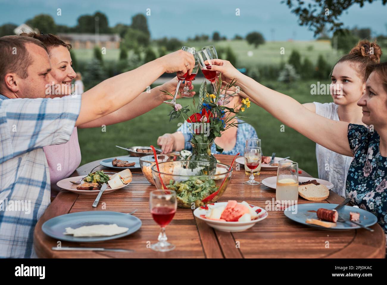 Family making toast during summer picnic outdoor dinner in a home garden. Close up of people holding wine glasses with red wine over table with food a Stock Photo