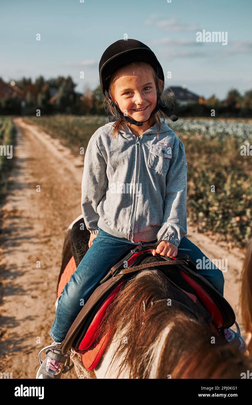 Little smiling girl learning horseback riding. 5-6 years old equestrian in helmet having fun riding a horse Stock Photo