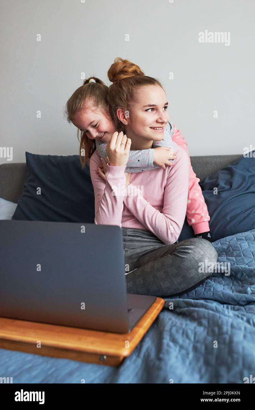 Teenage girl and her younger sister playing learning on laptop spending time together at home during COVID-19 quarantine. Girls sitting on bed in fron Stock Photo