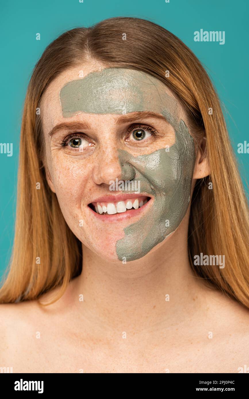 cheerful woman with red hair and clay mask on half of face smiling isolated on turquoise,stock image Stock Photo