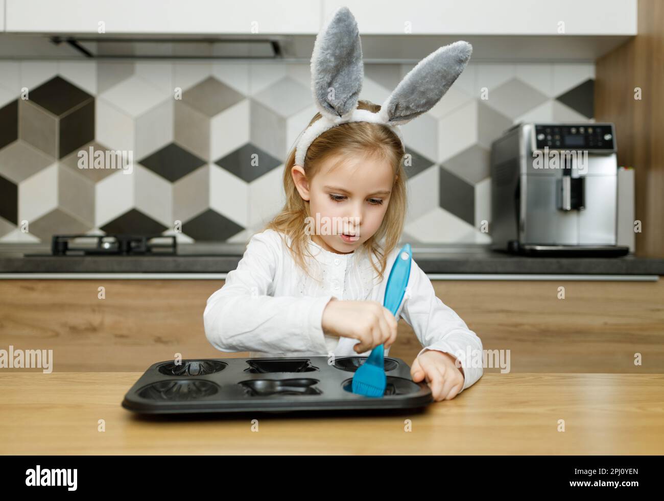 Serious child ittle girl in bunny ears lubricate baking dish with blue silicone brush with olive oil in modern wooden kitchen at home. Easter concept, Stock Photo