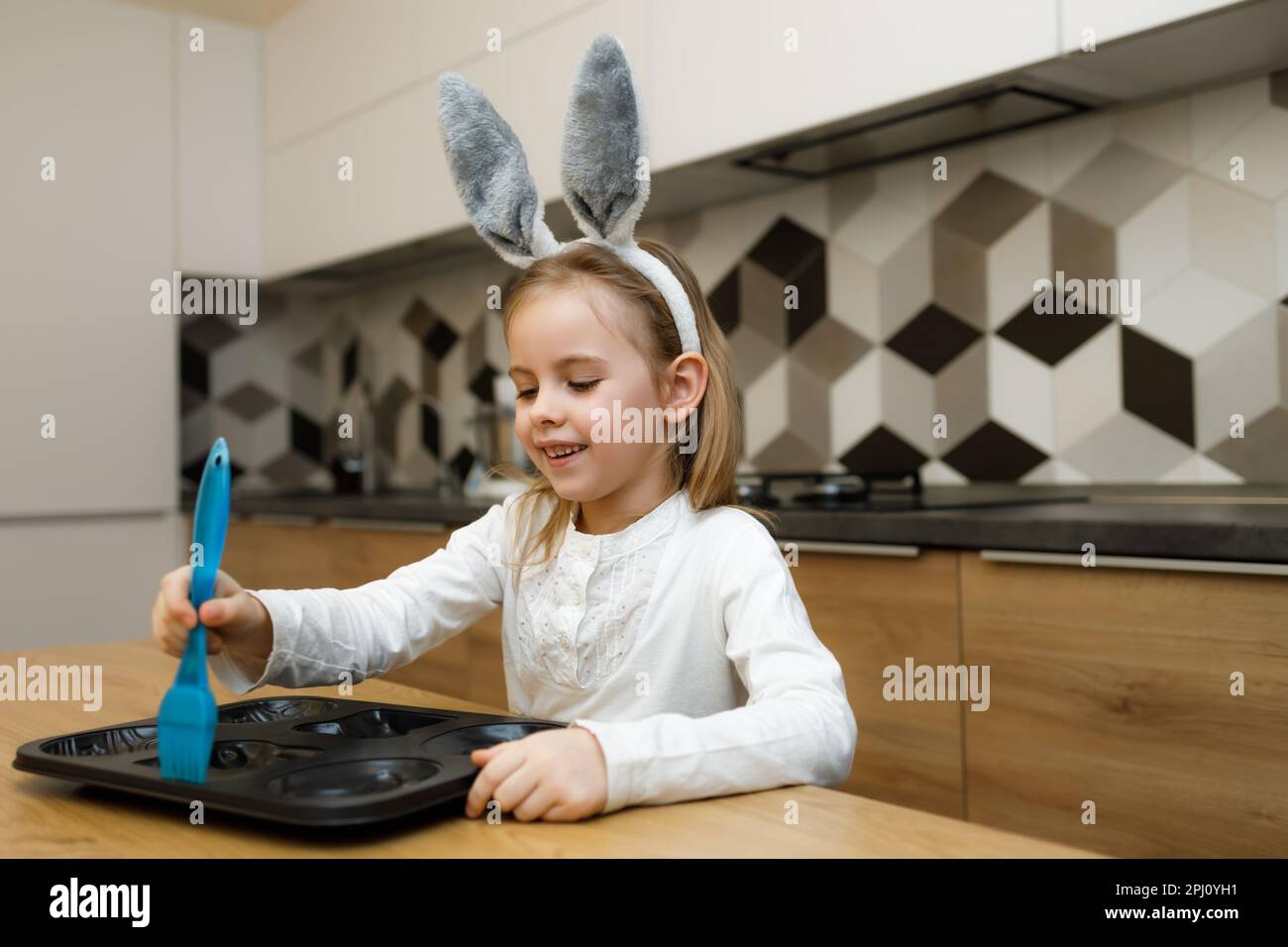 Little girl in bunny ears smiling and lubricate baking dish with silicone brush with olive oil in modern kitchen at home. Happy Easter concept, homema Stock Photo