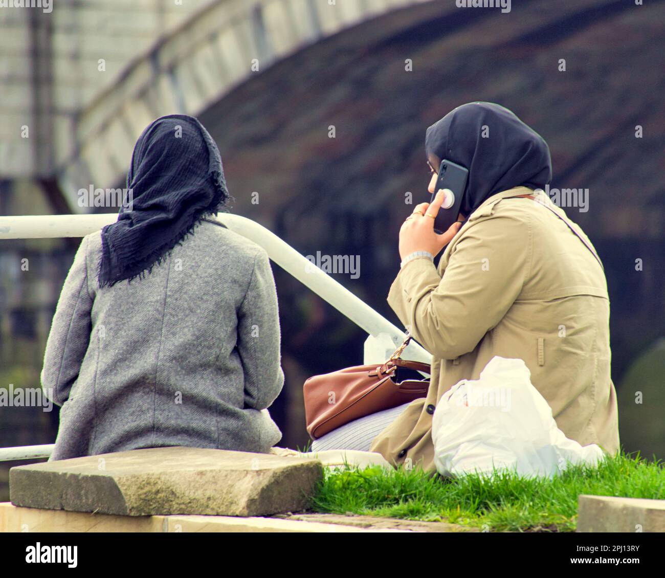 asian women with hijabs sitting beside river clyde on mobile phone Stock Photo