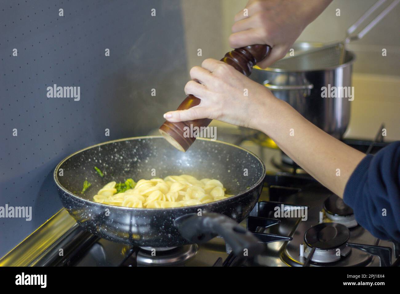 Image of a woman's hands seasoning freshly cooked pasta in a pan with a pepper mill. Italian culinary tradition. Stock Photo