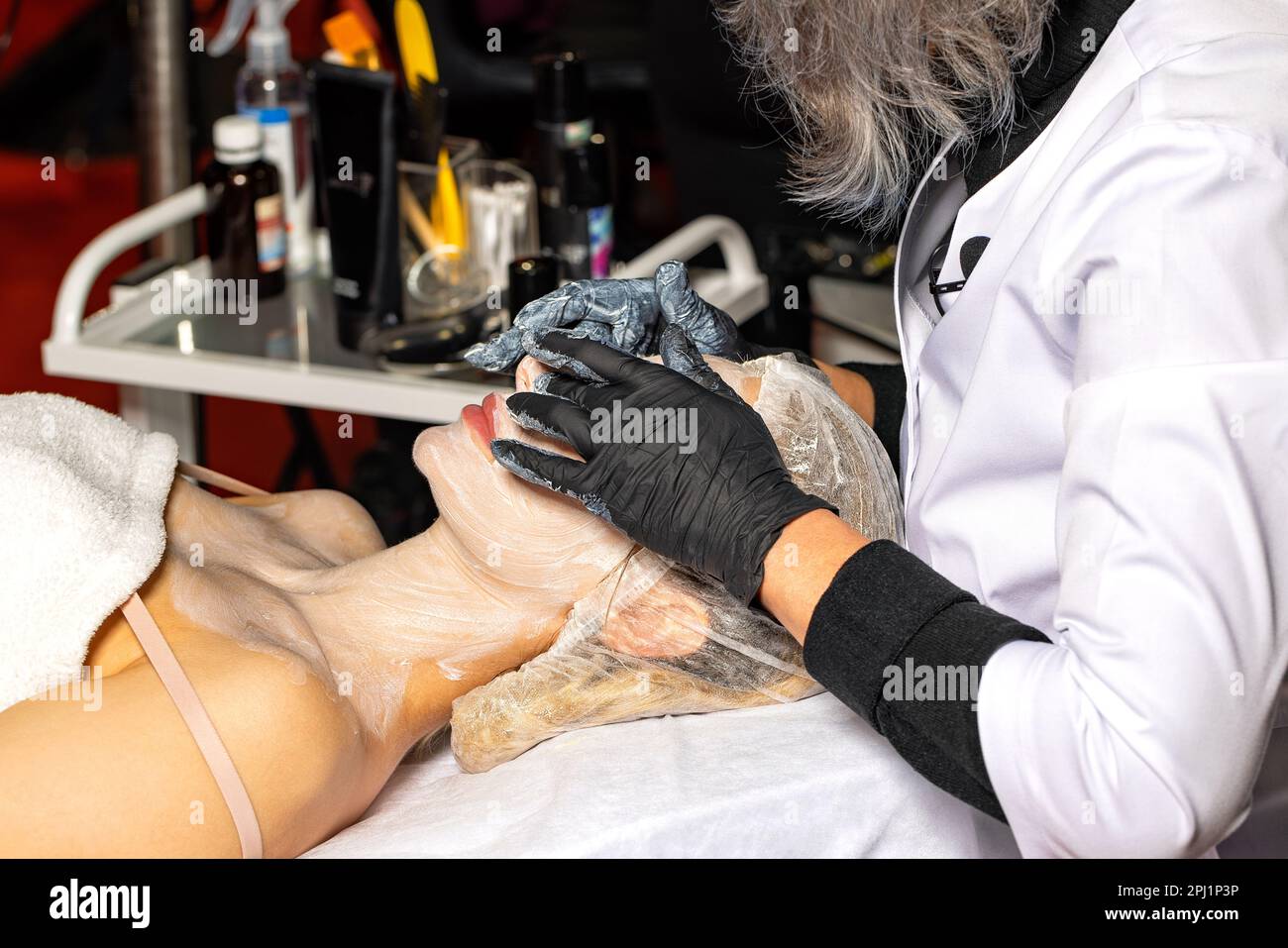 The hands of a beautician in black latex gloves do a facial cleansing procedure to a young woman. Stock Photo