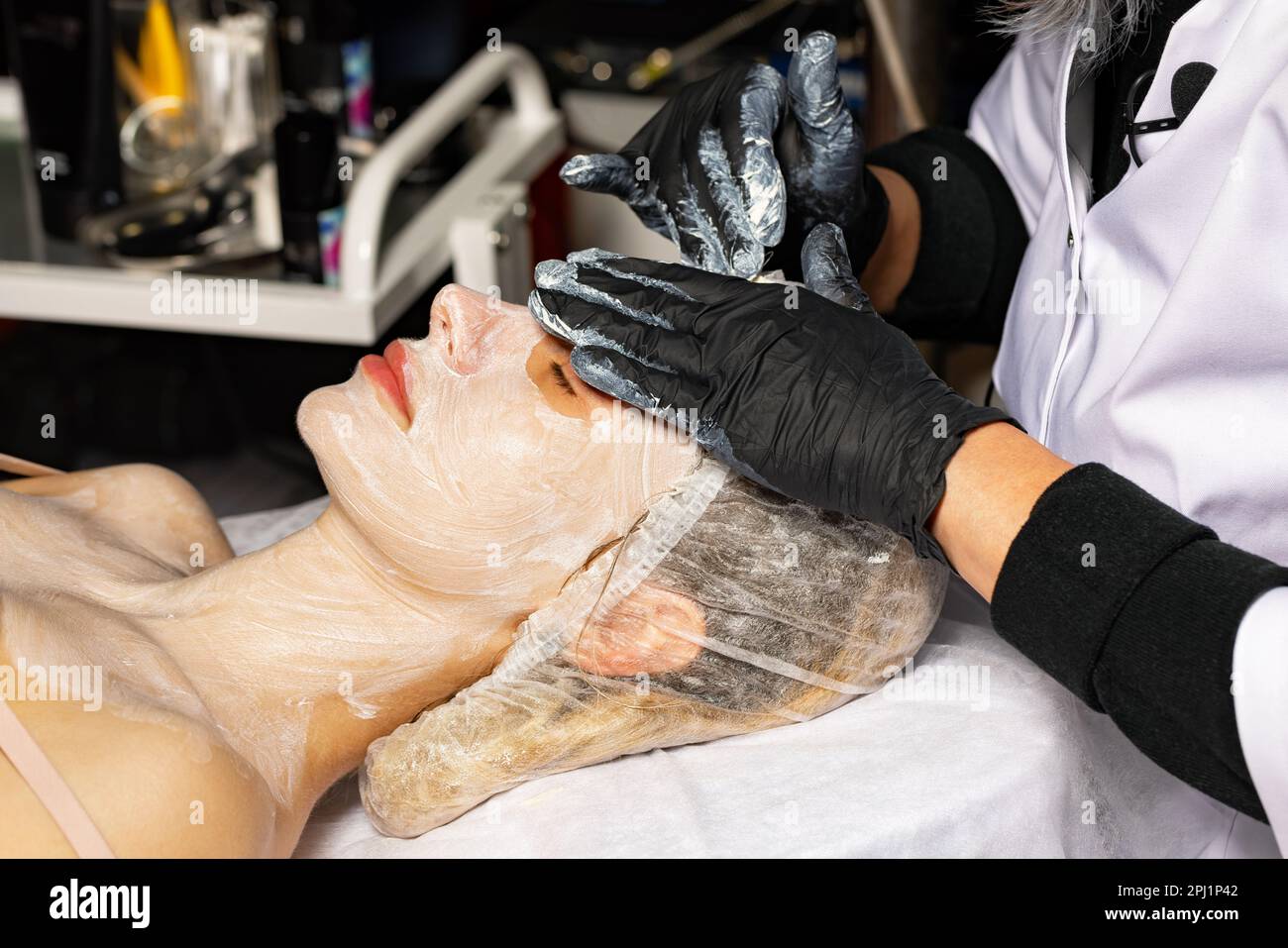 Professional beautician conducts hands in black latex gloves the procedure of cleaning the face of a young woman in a spa salon. Stock Photo