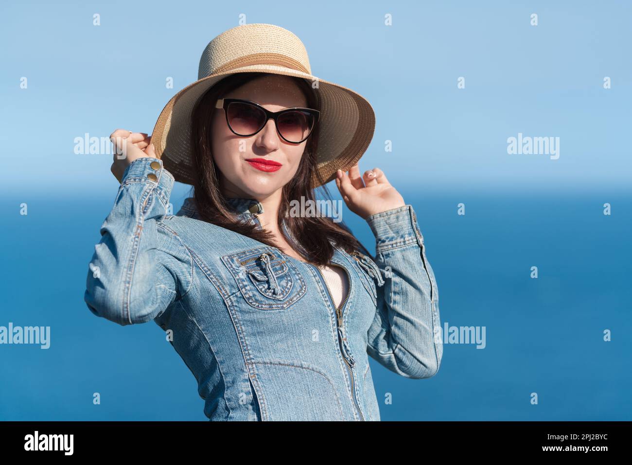 Caucasian ethnicity woman in sunglasses raised her hands and holding straw hat on her head. 40 year old hipster woman in denim jacket standing Stock Photo