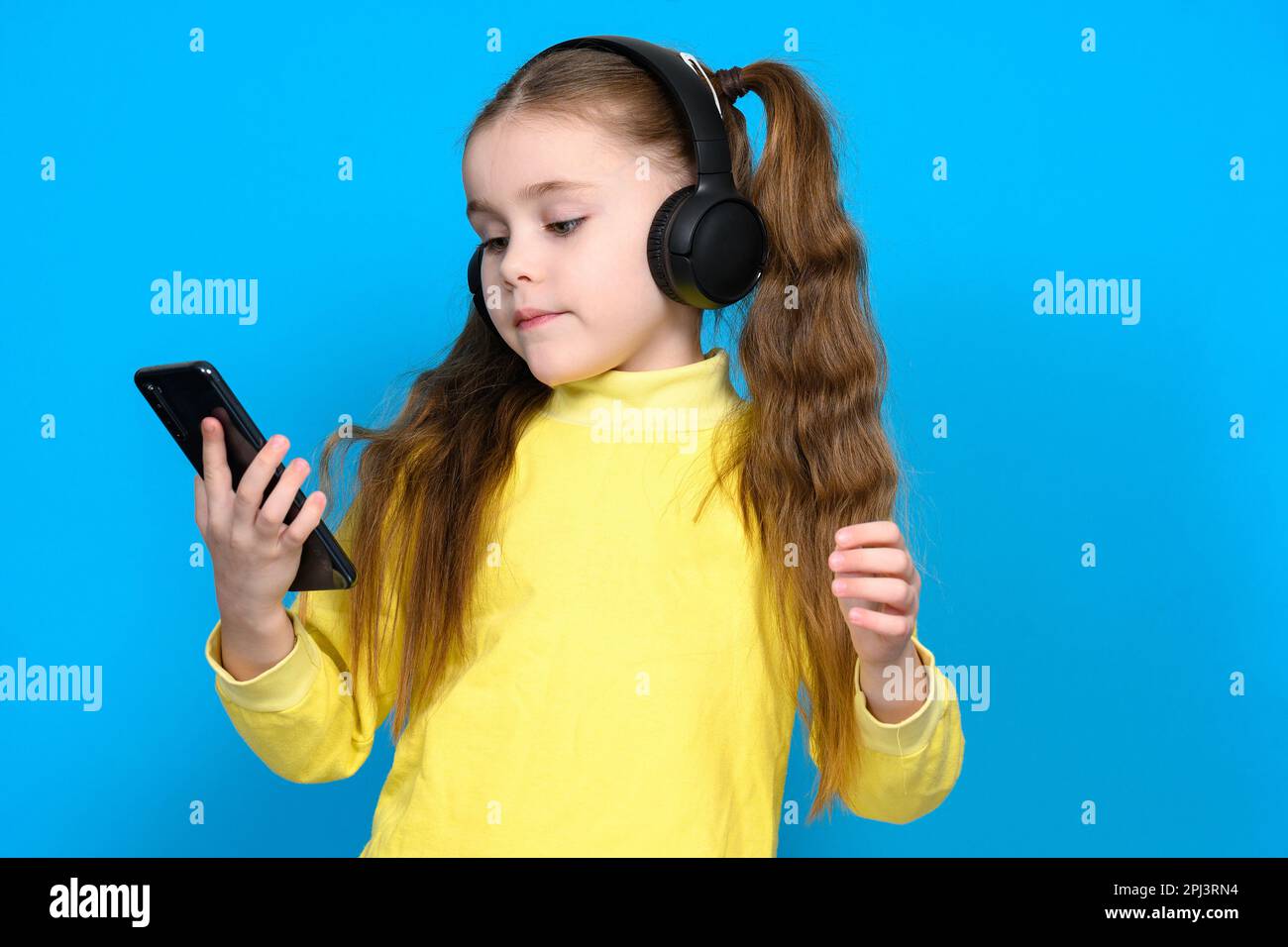 Portrait of a little girl with a phone and wireless headphones, a child in a yellow sweater on a blue background, a child dances when he hears music. Stock Photo
