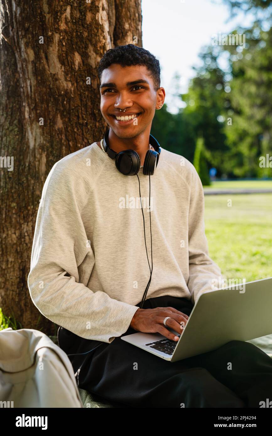Young black man smiling and using laptop while sitting under tree in park Stock Photo