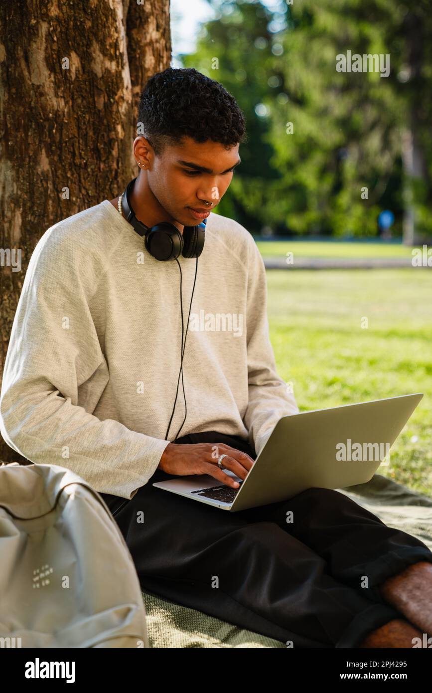 Young black man smiling and using laptop while sitting under tree in park Stock Photo