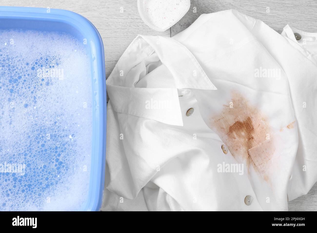 Dirty white shirt near basin with detergent foam in water on wooden
