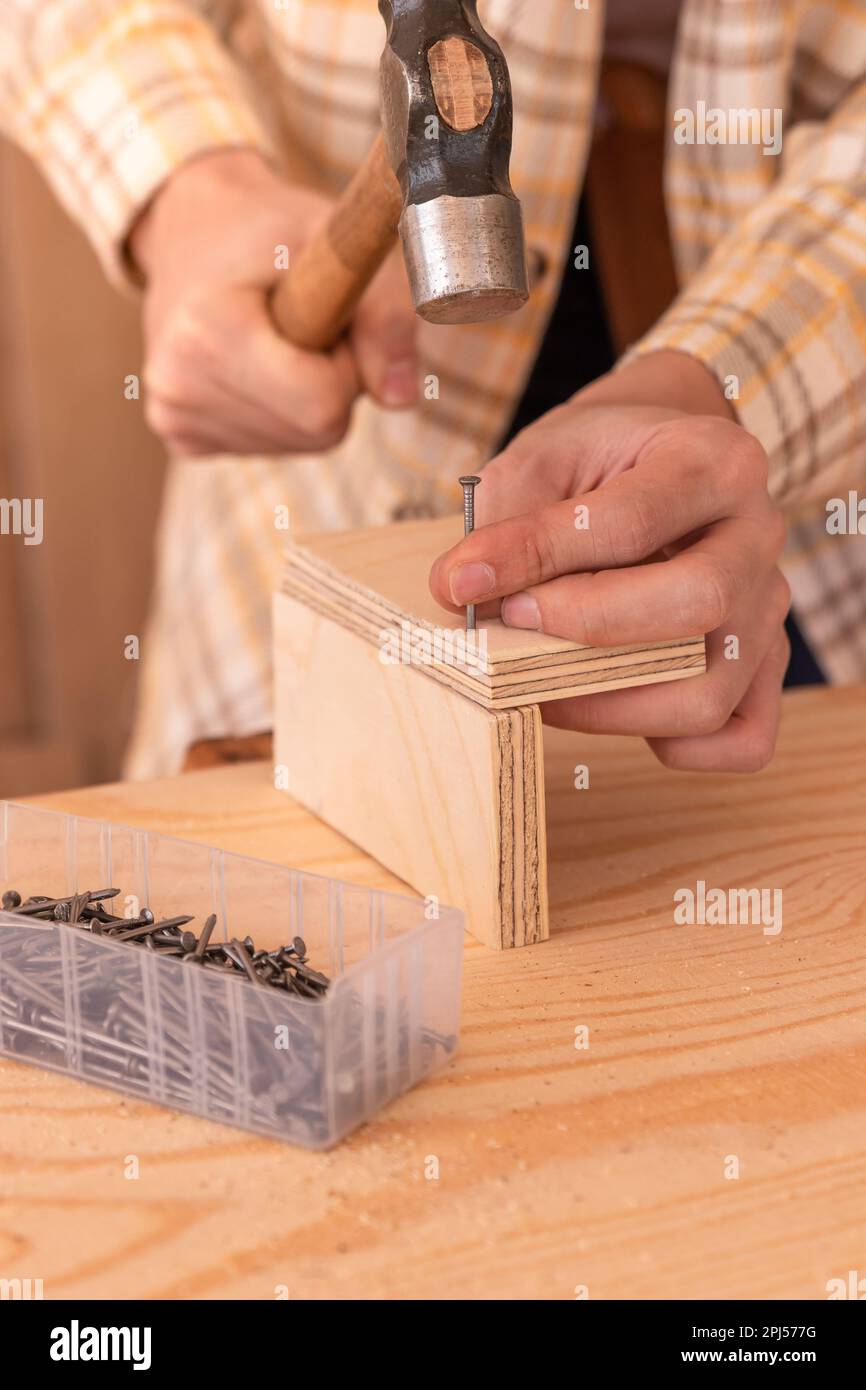 Crop anonymous male woodworker using hammer to drive nail into wooden board on workbench near box with nails while attaching pieces of furniture in wo Stock Photo