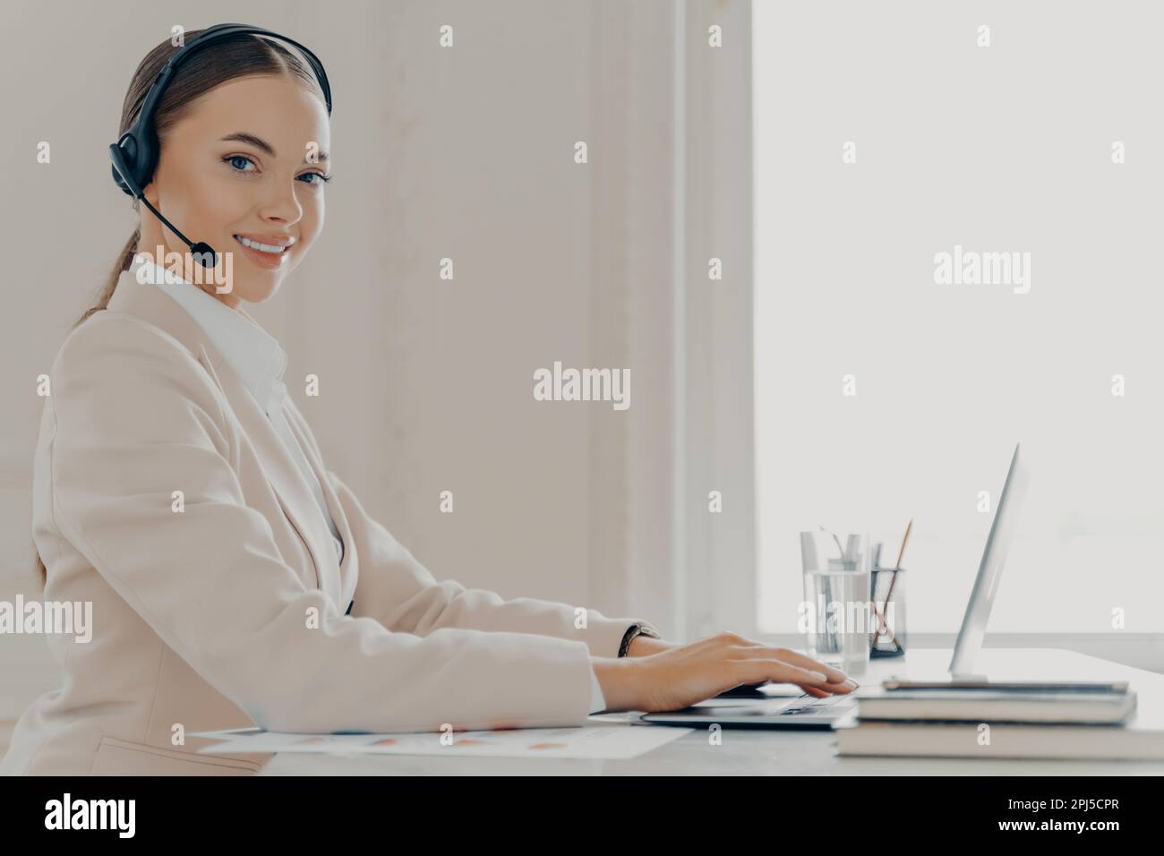 Sedi view of young female call center agent in headset with mic consulting client and using laptop while working in light offic, businesswoman wear in Stock Photo