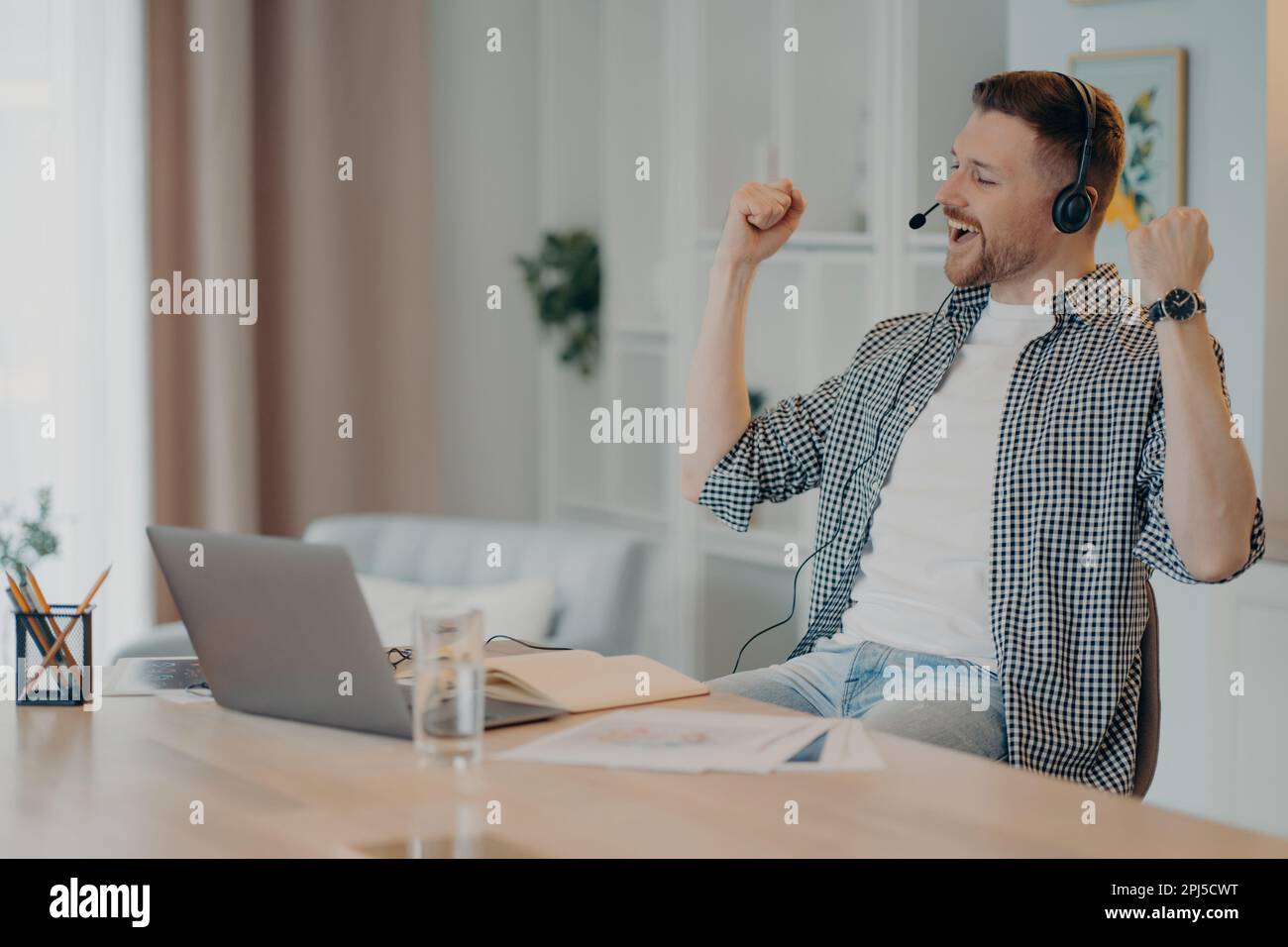 Happy successful man clenches fists celebrates triumph works online wears headset poses at desktop with laptop computer dressed casually. Overjoyed ma Stock Photo