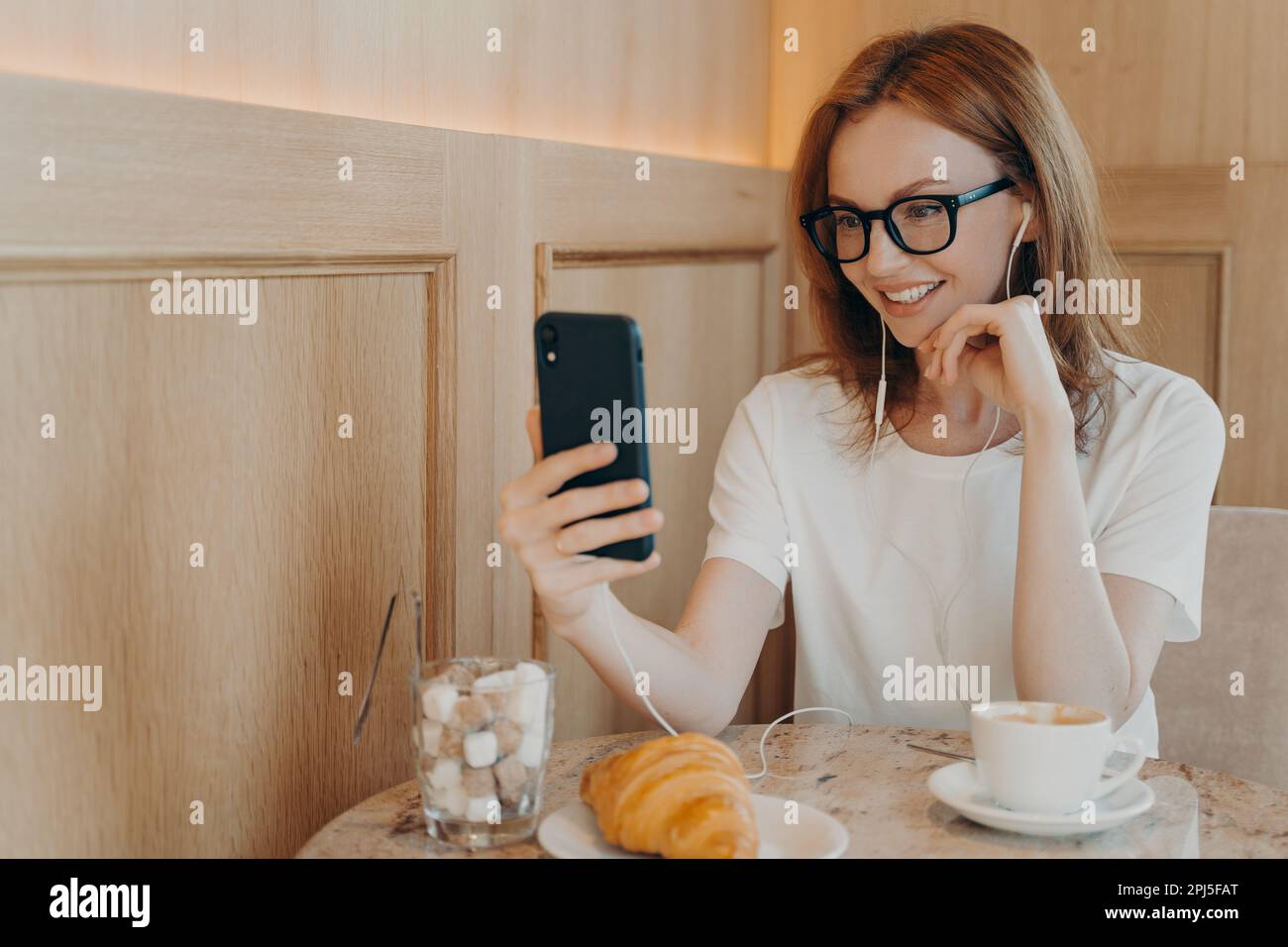 Indoor shot of cheerful redhead young woman uses mobile phone and earphones for online conversation dressed casually sits in cozy coffee shop has frie Stock Photo