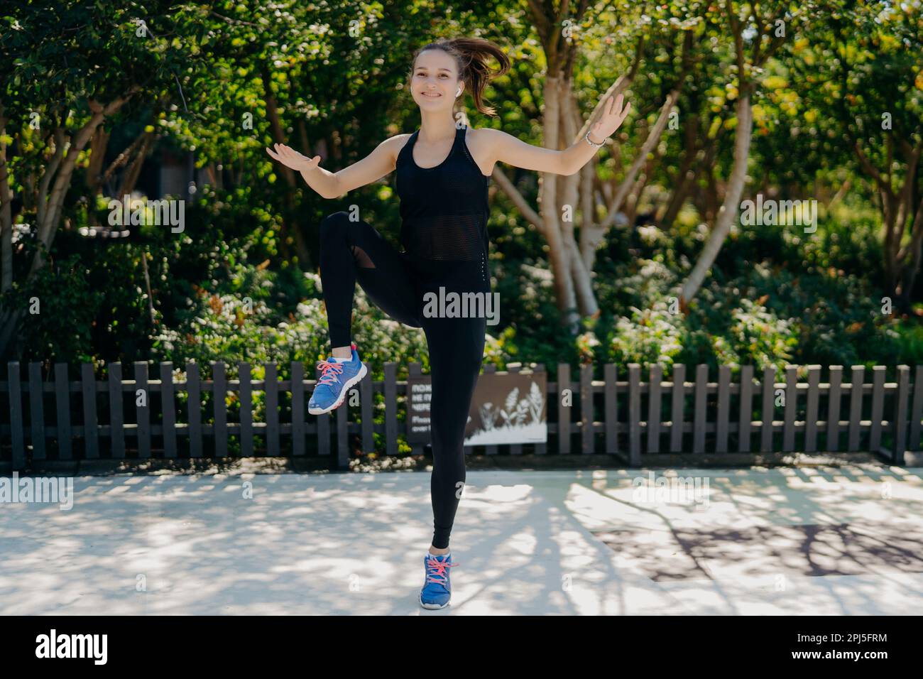 Outdoor shot of athletic motivated woman jumps high does physical exercises keeps hands and legs raised dressed in black sport clothes being in good m Stock Photo