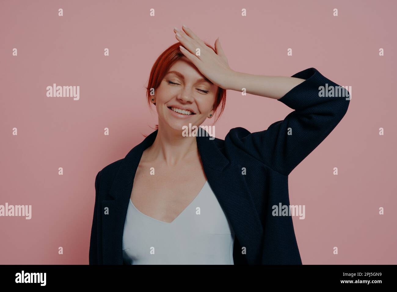 Beautiful laughing carefree red-haired woman isolated on pink studio background keeping hand on forehead and eyes closed, smiling broadly and feeling Stock Photo