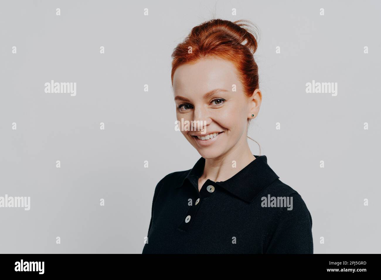 Close up portrait of young beautiful redhead female with natural makeup and beaming smile looking at camera, posing isolated over grey background, att Stock Photo
