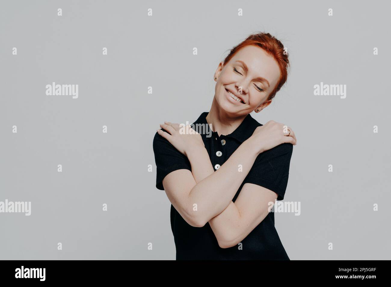 Happy red-haired woman with closed eyes dressed casually hugging herself gently and tilting head, pretty ginger female being in romantic mood while st Stock Photo