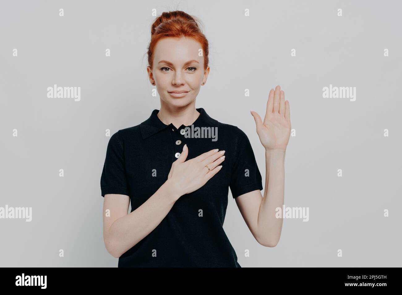 Swear to tell only truth. Studio shot of serious red haired female wearing black t-shirt making promise, swearing with hand on chest while standing ag Stock Photo