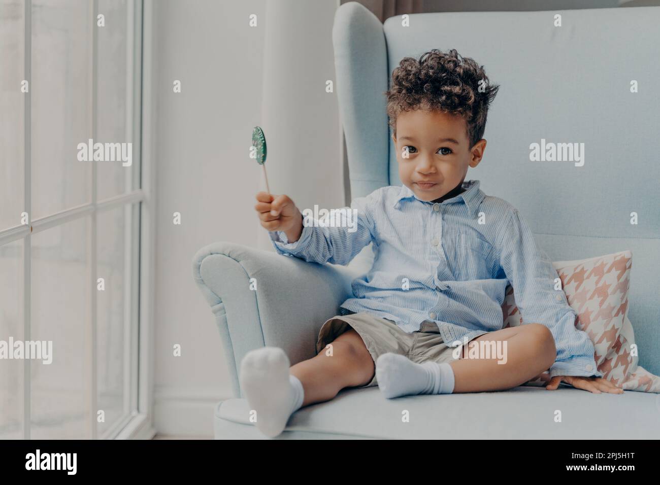 Portrait of cute adorable little mulatto boy with big eyes holding round blue sweet and tasty lollipop and looking at camera, casually dressed sitting Stock Photo