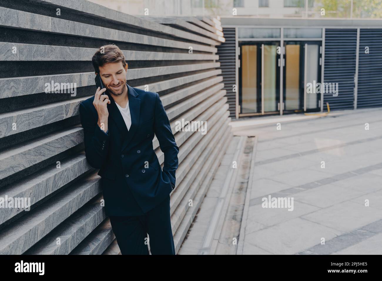 CEO male in smart casual having pleasing phone talk standing outside of office center entrance, leans on marbale striped facing outer wall, having cov Stock Photo