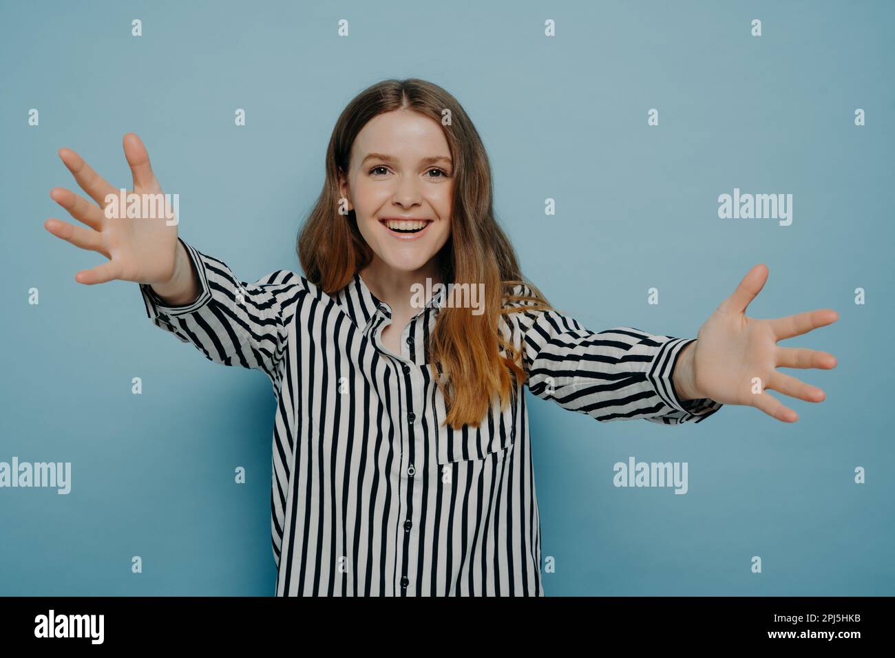 Portrait of loving young woman offering hug, smiling and pulling hands towards camera while standing isolated on blue background, cheerful teenage gir Stock Photo