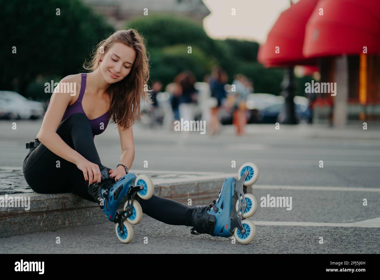 Outdoor shot of young sporty woman in sportswears puts on roller blades poses on road going to ride rollers during sunny day ties shoelaces has good m Stock Photo