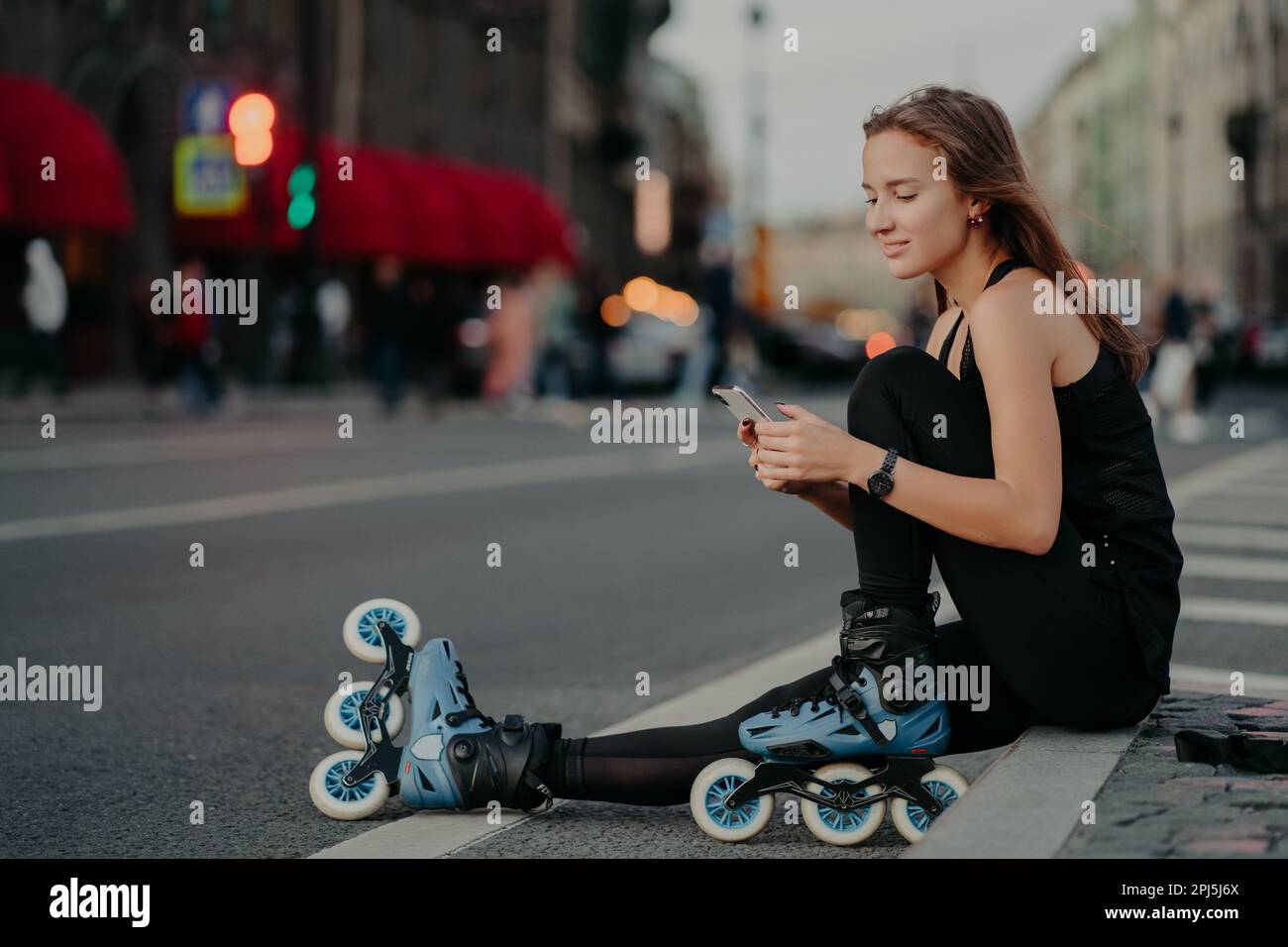 Outdoor shot of sporty active young woman dressed in active wear wears rollerblades checks message content on smartphone poses against blurred city ba Stock Photo