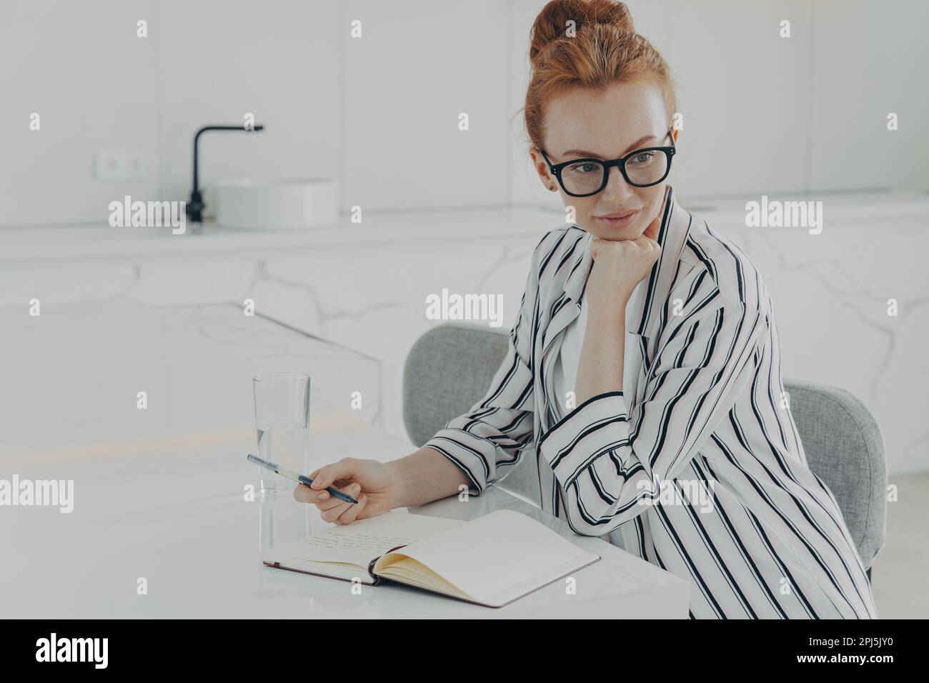 Young concentrated ginger woman in eyewear dressed casually thinking about new day while sitting at table in kitchen with transparent glass of water, Stock Photo