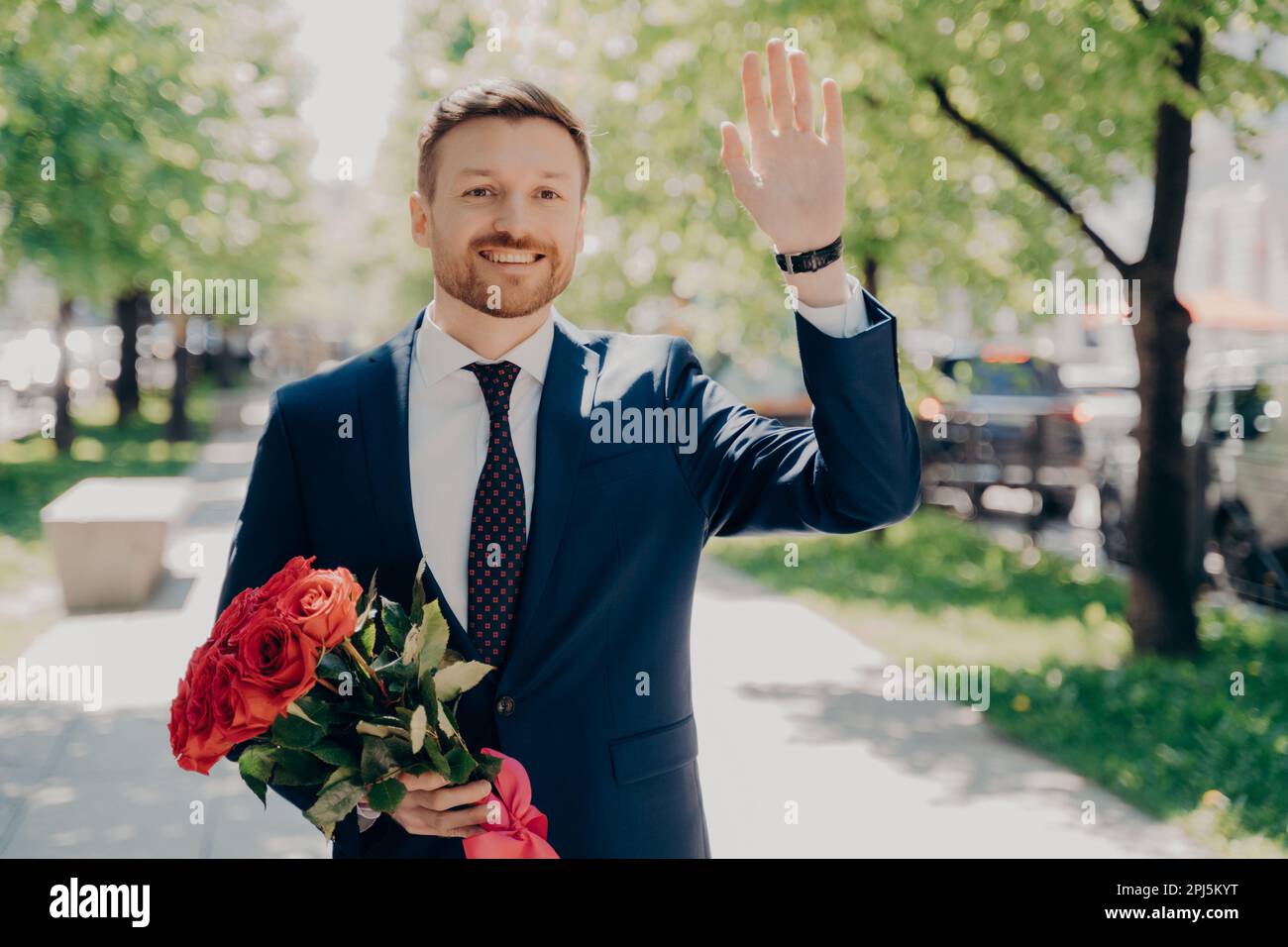 Happy handsome young man in blue suit holding fresh bouquet of red ...
