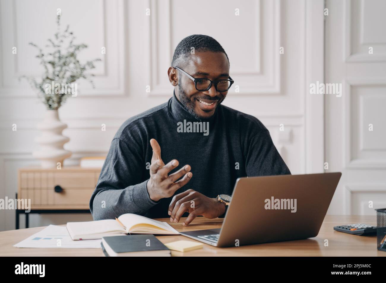 African-American young businessman in glasses having conference online with employes via video call broadly smiling while looking at laptop screen ges Stock Photo