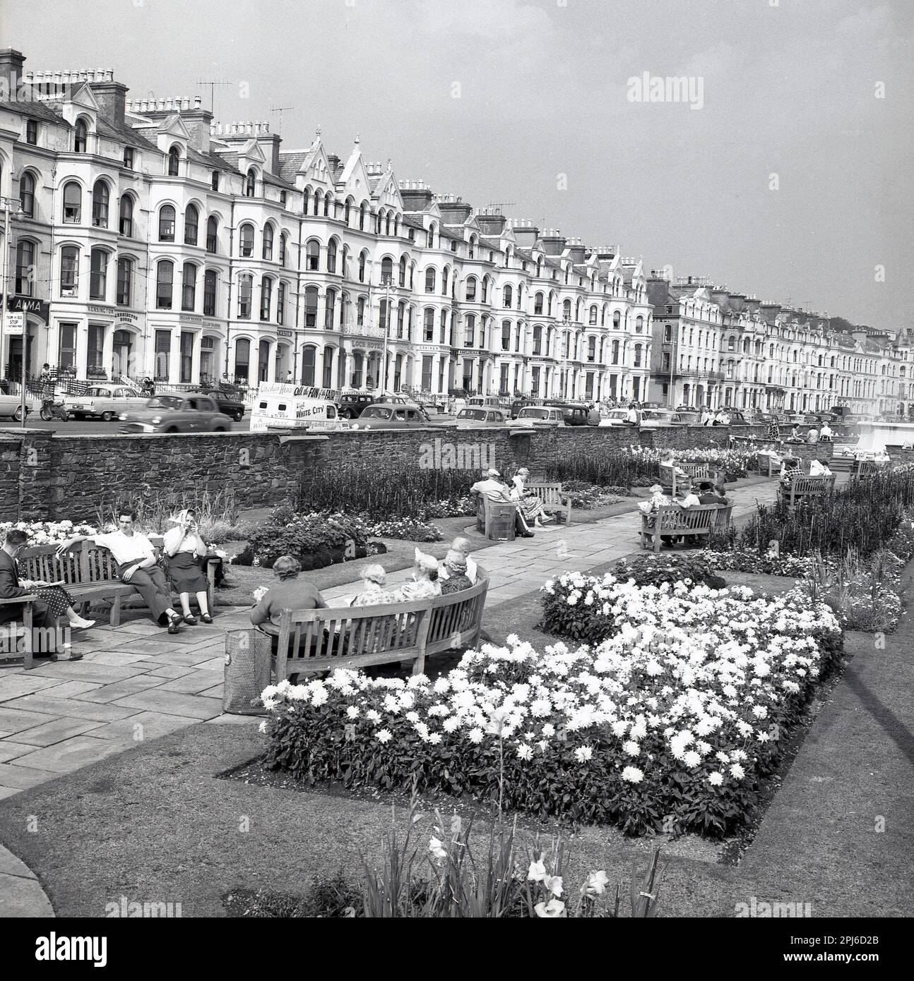 1950s, historical view from this era of people siting on benches at the seafront gardens at Brighton, England, UK. The victorian terraced was the home of many hotels catering for the holidaymakers. A van of the era is parked in the street advertising, Onchan Head...all the Fun of the Fair...Visit White City. Stock Photo