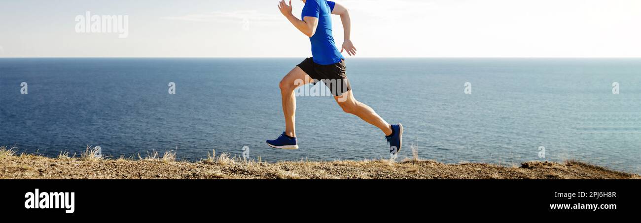 male runner running along seashore trail with dry grass on blue sky background, cross-country running race Stock Photo