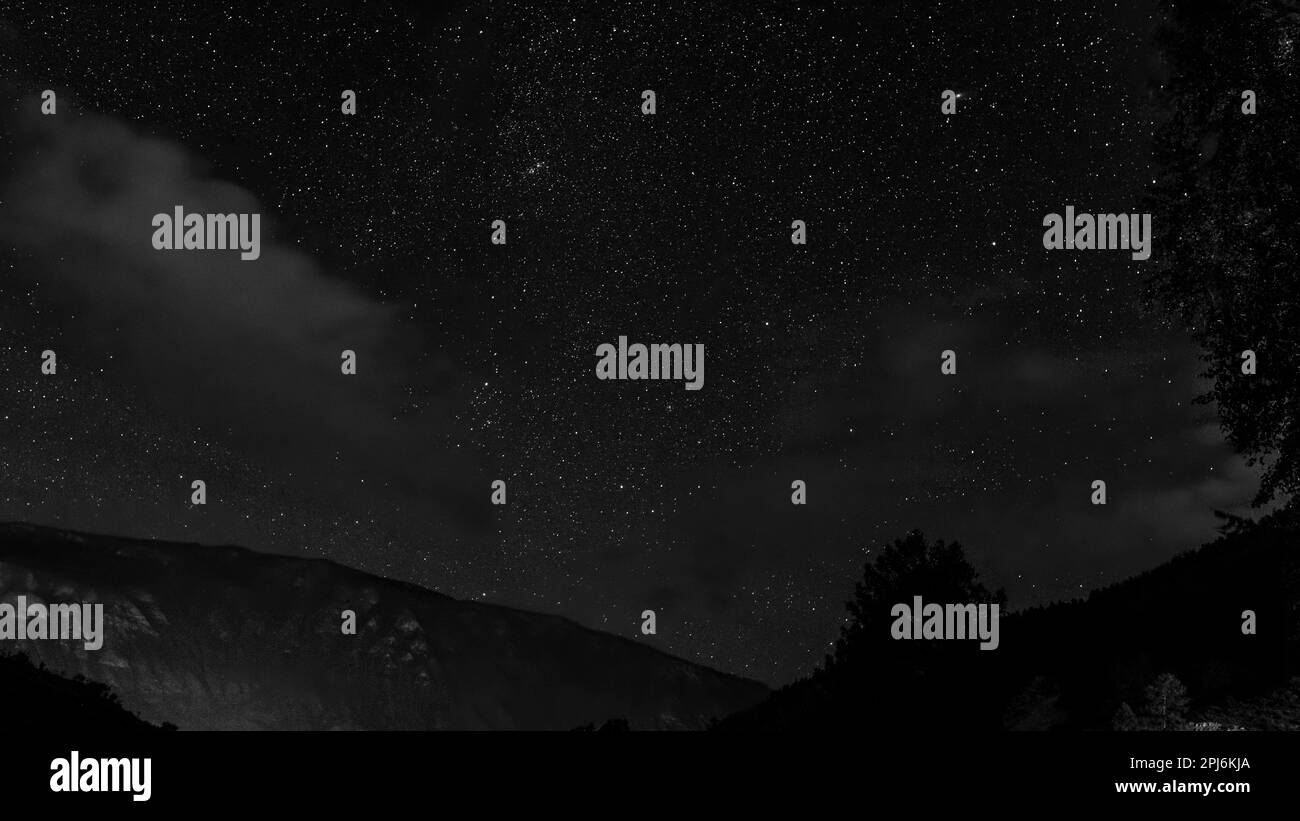 Black and white photo of a starry sky at night over a mountain and a forest with clouds in the trees in the Altai in Siberia. Stock Photo