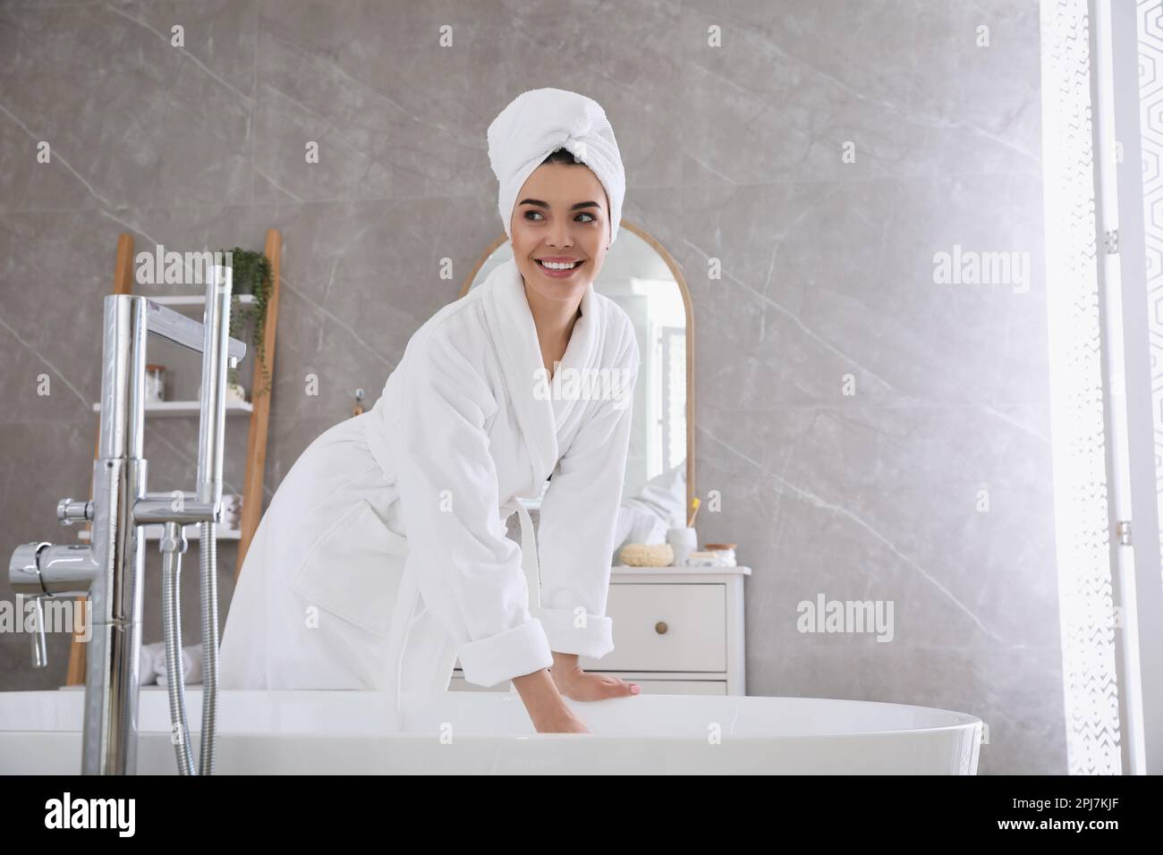 Beautiful young woman in soft white robe near bathtub at home Stock