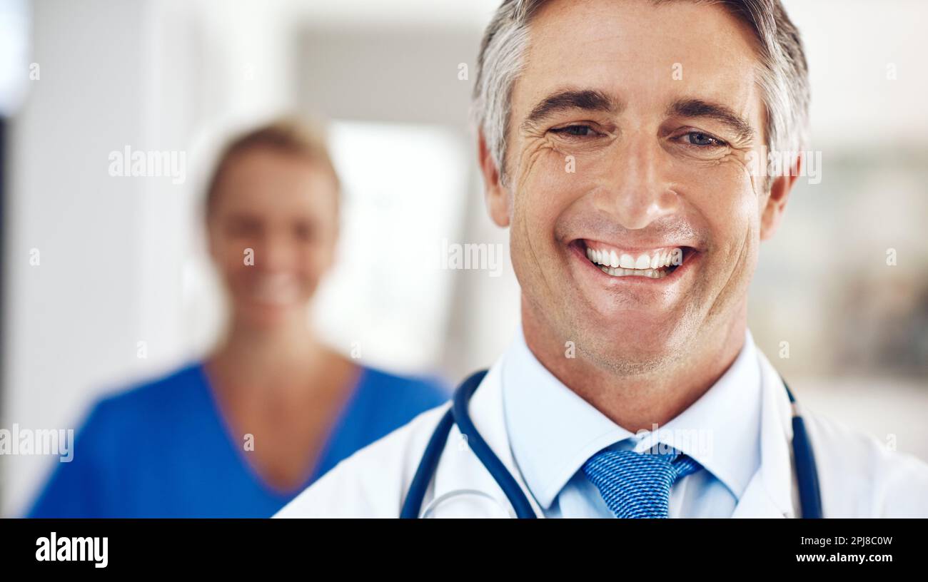 Greeting his patients with a smile. Portrait of a handsome mature male doctor standing in the hospital with a female colleague in a the background. Stock Photo