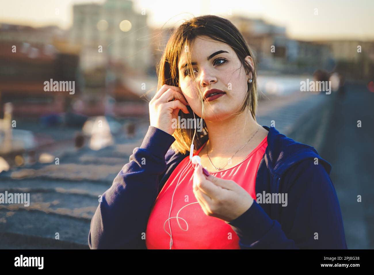 A young plus-size woman takes a break during her outdoor workout. She is wearing a jogging suit and is seen wearing wired earphones. The background sh Stock Photo