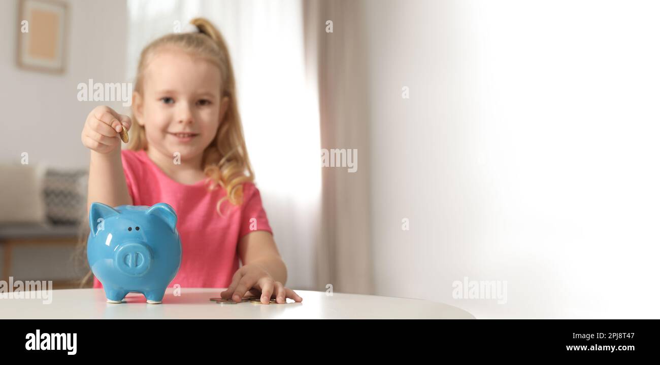 Cute girl putting coin into piggy bank at table in living room, space ...