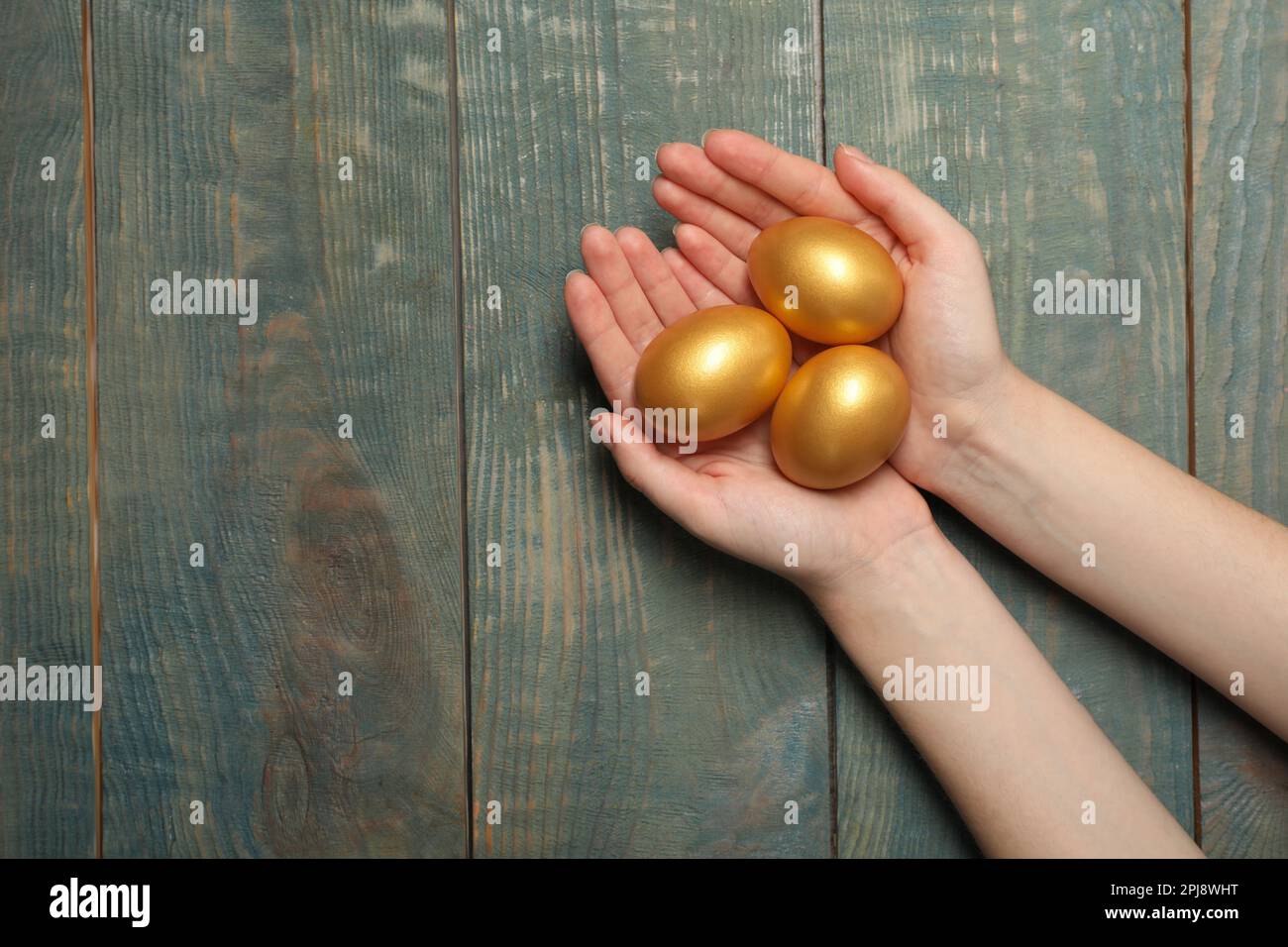 Woman holding shiny golden eggs at light blue wooden table, top view. Space for text Stock Photo