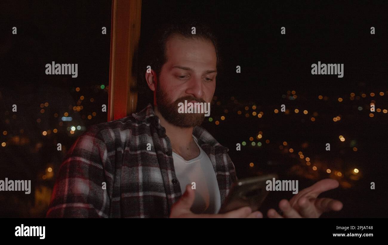 Handsome Man with Long Hair and Beard in Checked Shirt Uses Messaging or Apps on Smartphone in a Stunning Home with a View Stock Photo